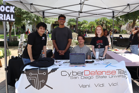 Four students tabling at a table with a computer and a tablecloth that says Cyber Defense Team